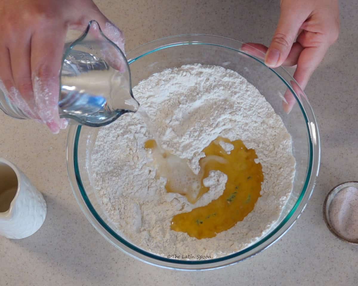 A glass bowl filled with flour is held in one hand while the other pours a little water from a pitcher, with a small bowl of salt and oil beside it.