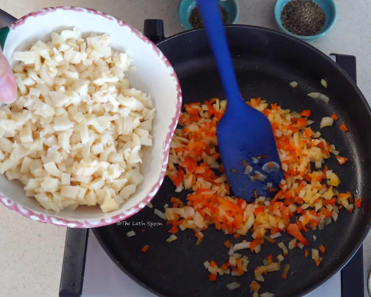 A skillet with a spatula, diced red bell pepper, and onion, while chopped hearts of palm are poured in from a cup.
