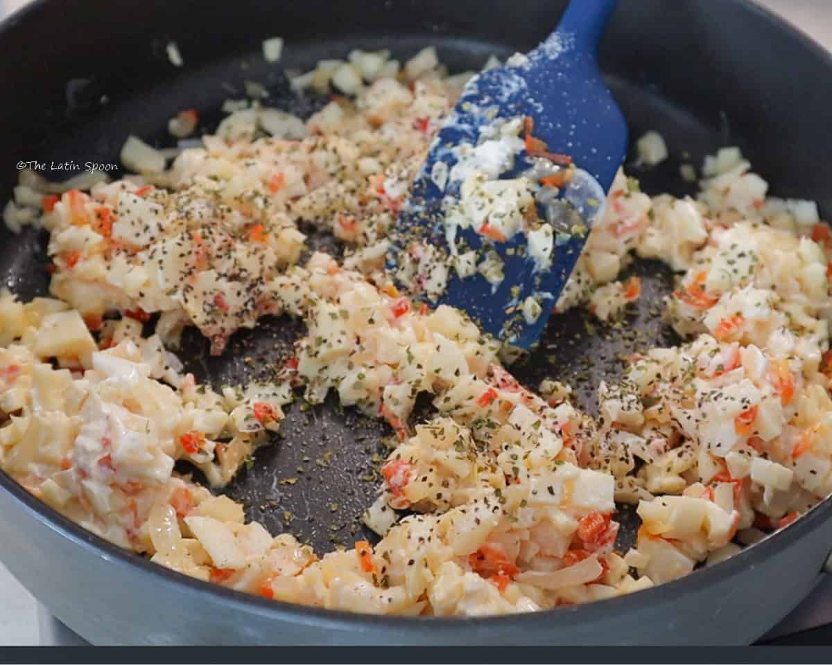 A skillet containing a mixture of diced onion, red bell pepper, and chopped hearts of palm being stirred with a spatula and sprinkled with ground black pepper.