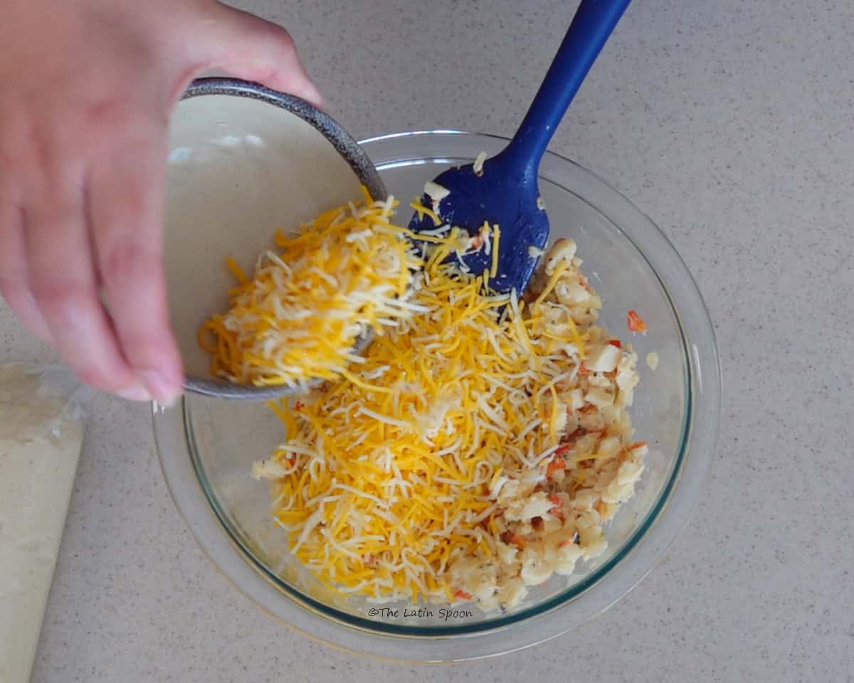 A glass bowl containing the heart of palm mixture, with grated cheese being poured in from a small cup and a spatula resting inside the bowl.
