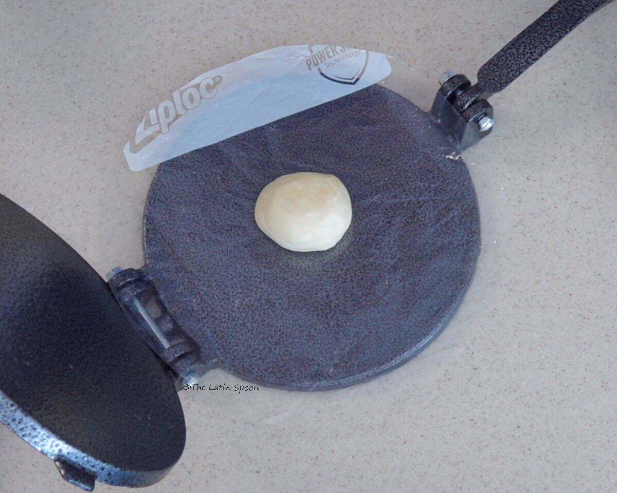 A tortilla press lined with two sheets of plastic, with a ball of flour dough placed in the center.