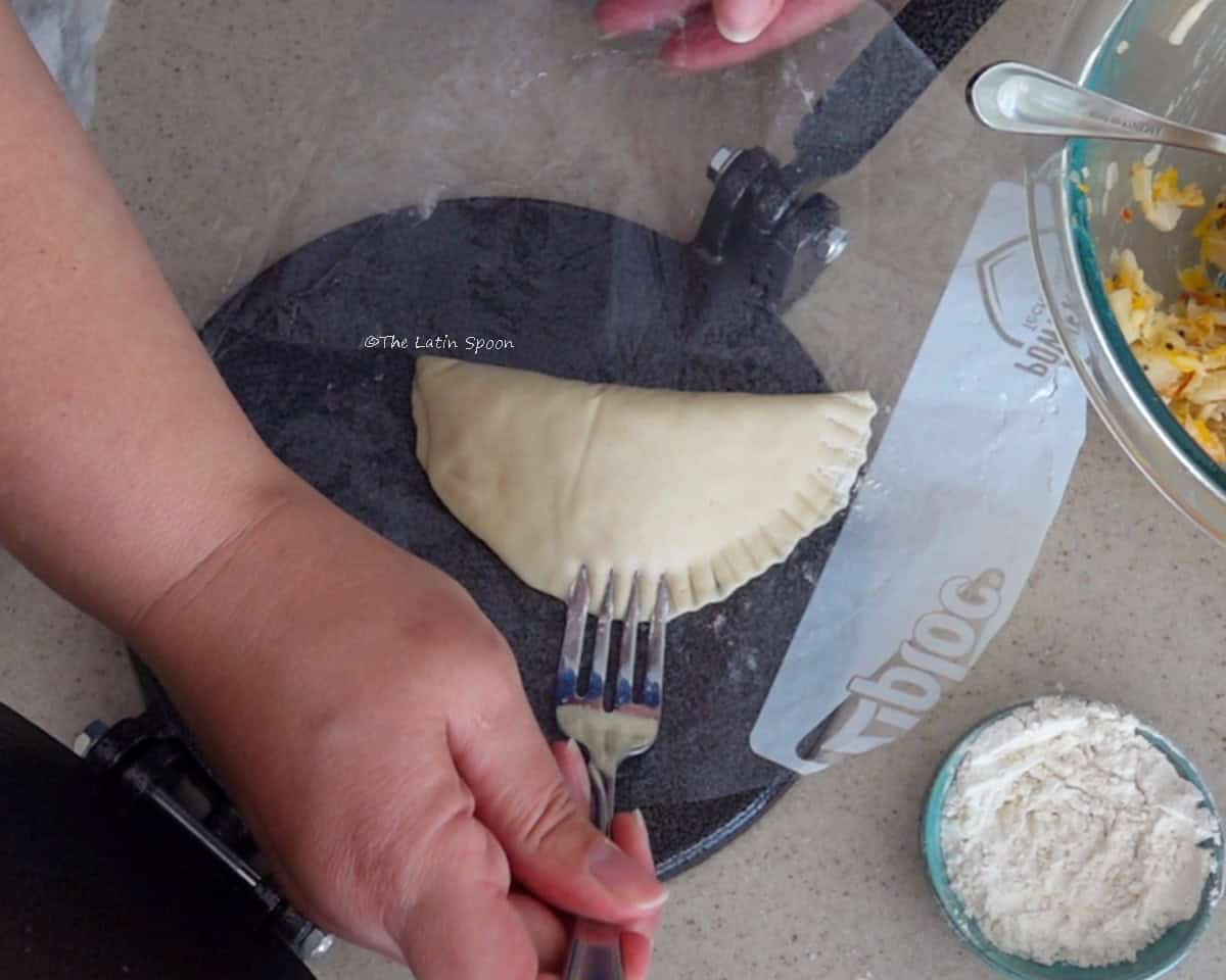 A hand uses a fork to crimp the edges of an empanada while it remains on the plastic-lined tortilla press.