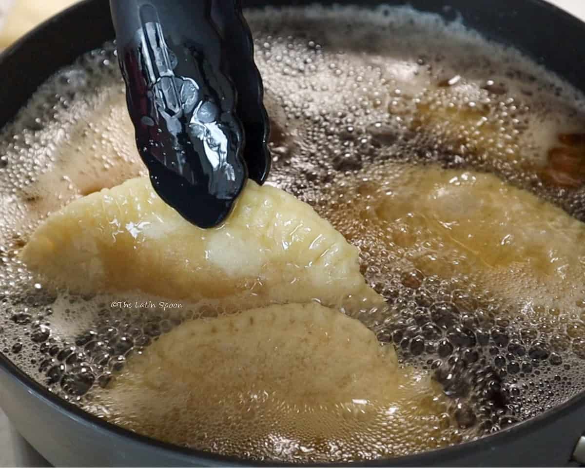 A skillet filled with hot oil and empanadas, with a pair of tongs turning them as they fry.
