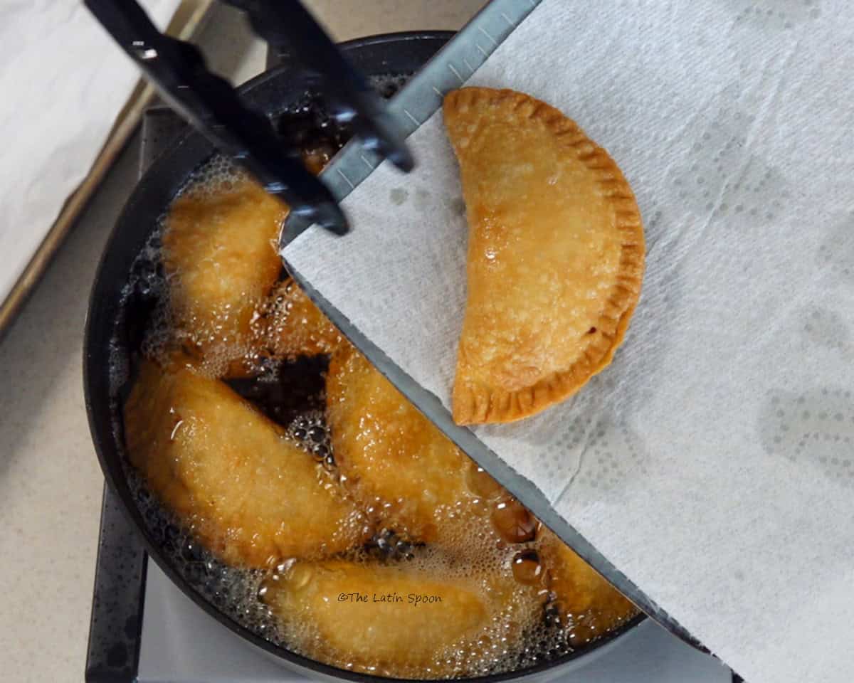 Fried empanadas being removed from the hot oil with tongs and placed on a paper towel-lined tray.