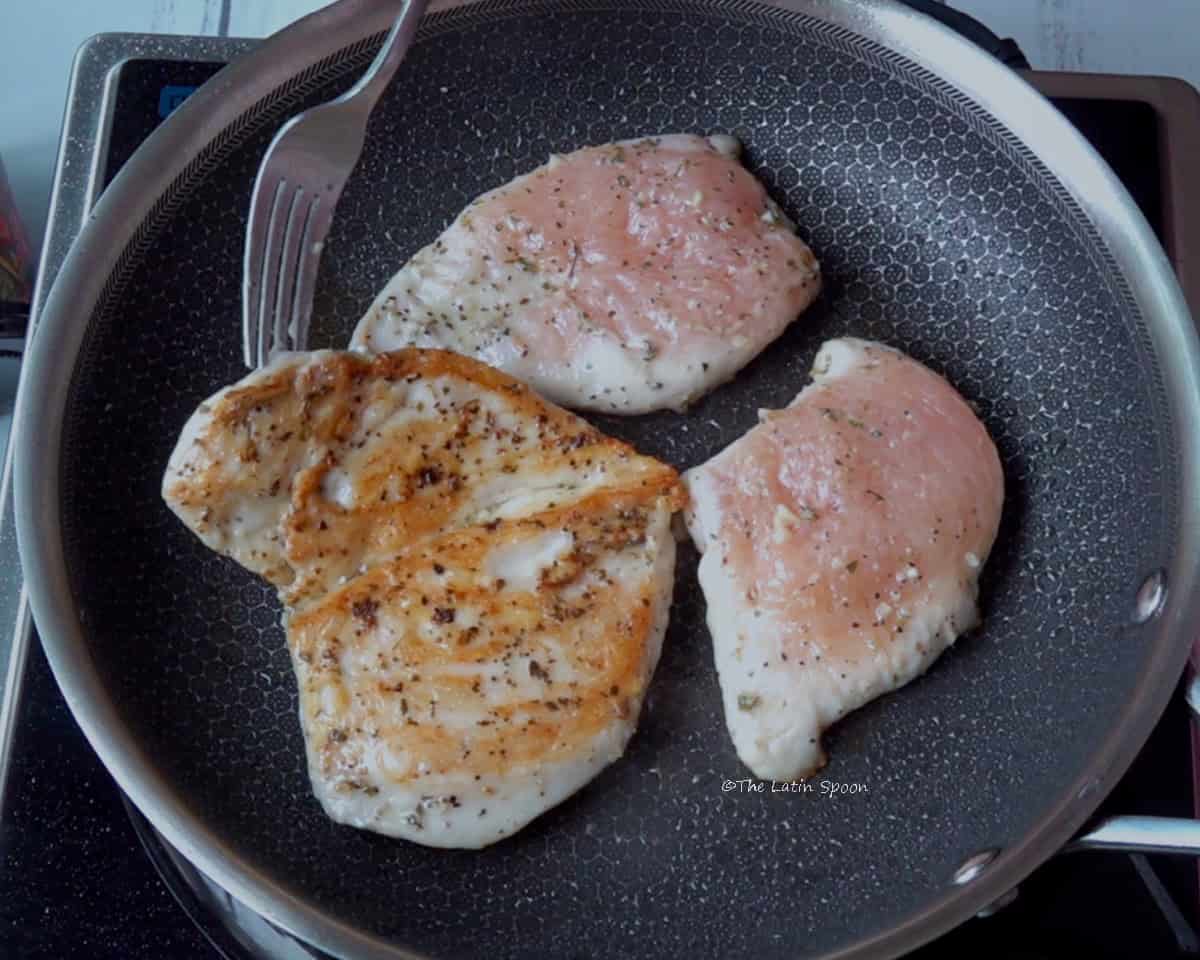 A stainless steel skillet in which three pieces of chicken are cooking. One of the pieces has been turned over, showing its golden-brown sear, while the other two pieces still appear raw.