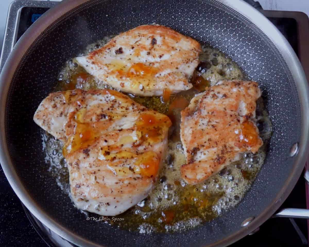 Four pieces of chicken in a stainless steel skillet, cooking with pineaplle habanero jam, showing the jam’s bright, vibrant color.