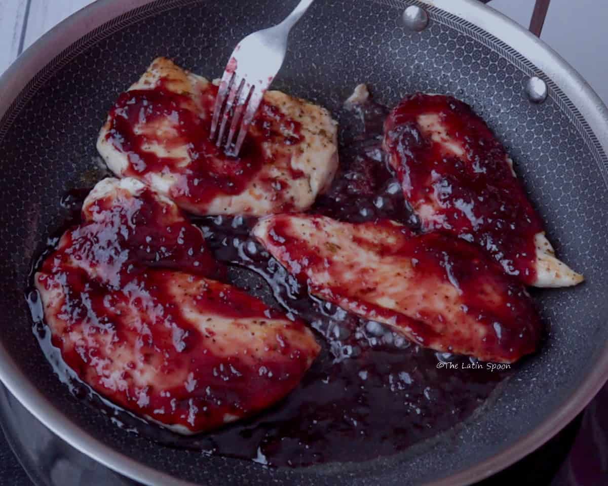 Four pieces of chicken in a stainless steel skillet, cooking with cherry jam, showing the jam’s bright, vibrant color.