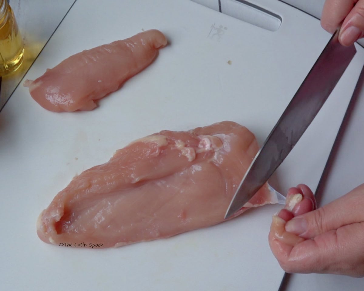 A white cutting board with a chicken breast on top. The left hand holds a piece of chicken while the right hand trims the excess fat from the breast.
