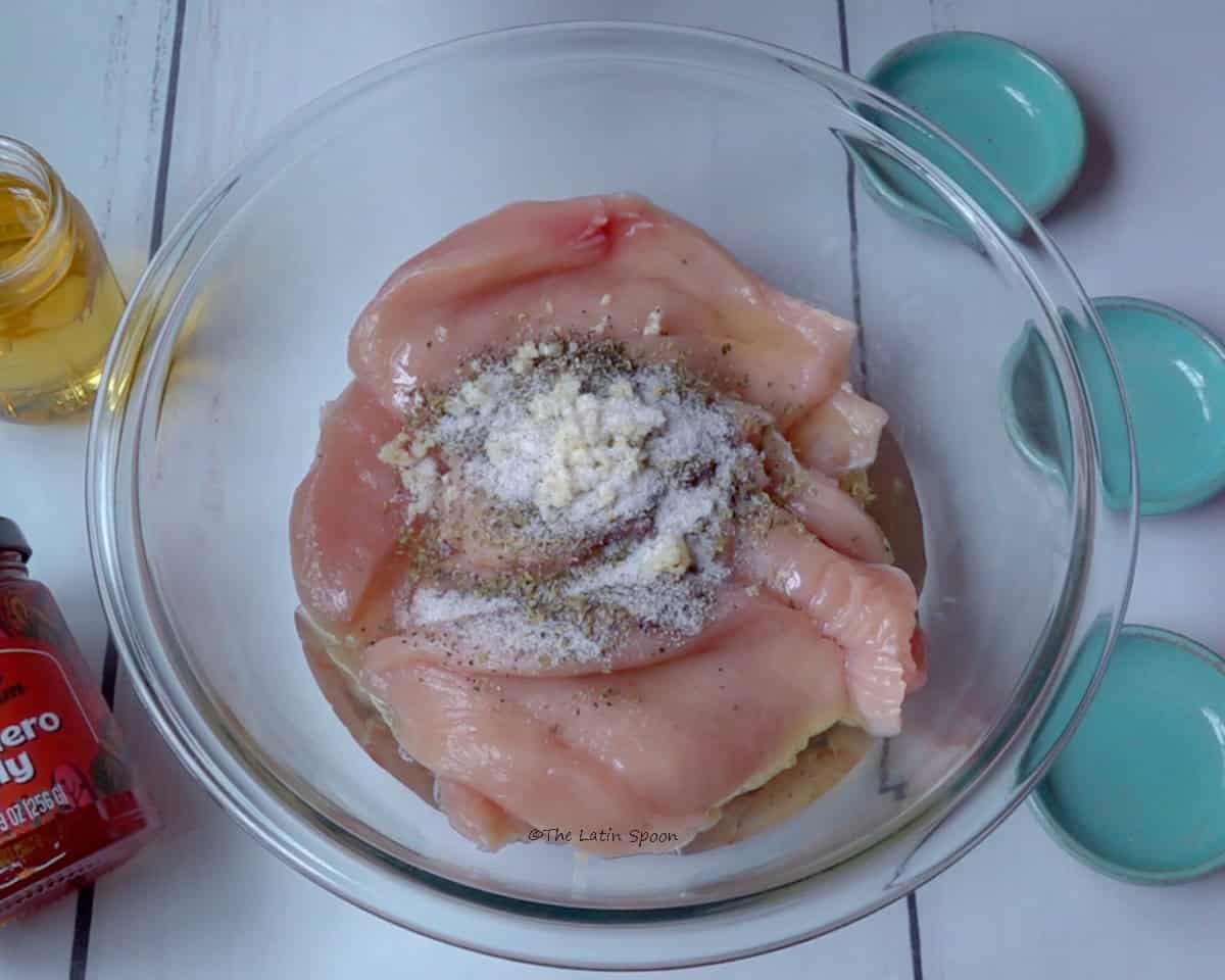 A transparent glass bowl containing small pieces of raw chicken. On top, you can see the oil and the spices used to marinate the chicken.