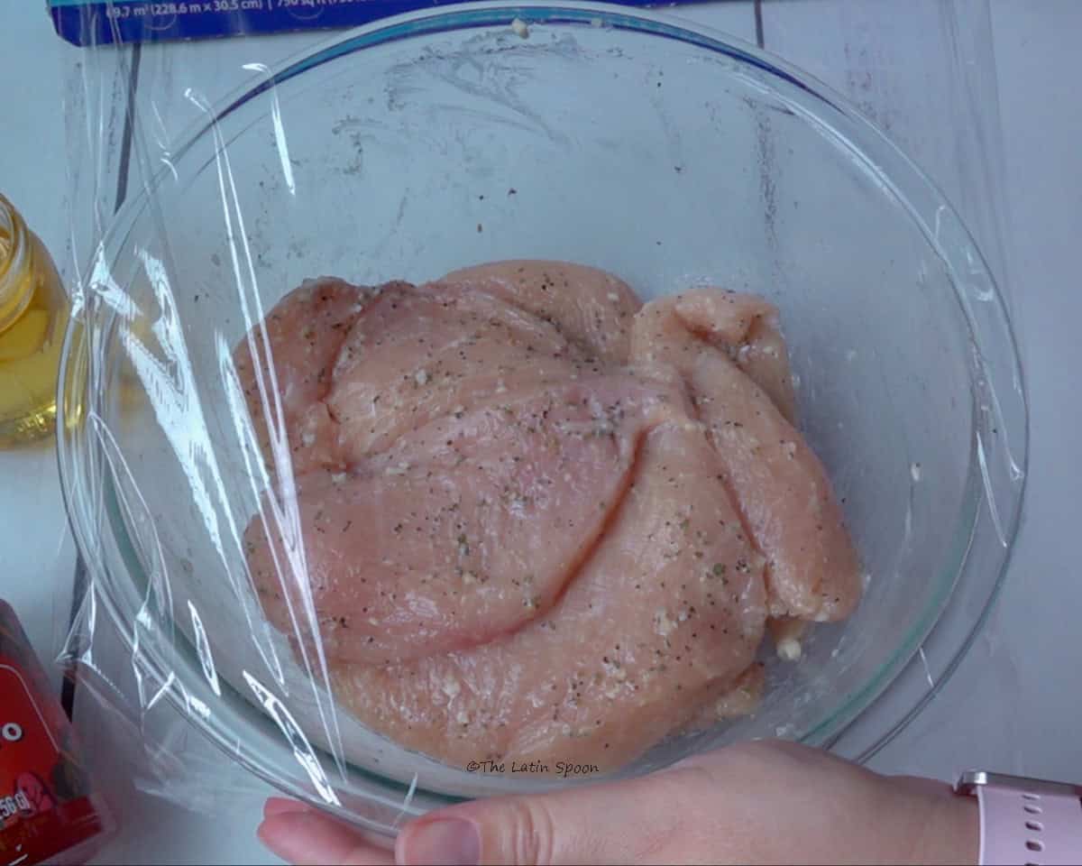 A transparent glass bowl containing the marinated chicken. A thin layer of plastic wrap is placed over the bowl to cover the chicken before refrigerating it.