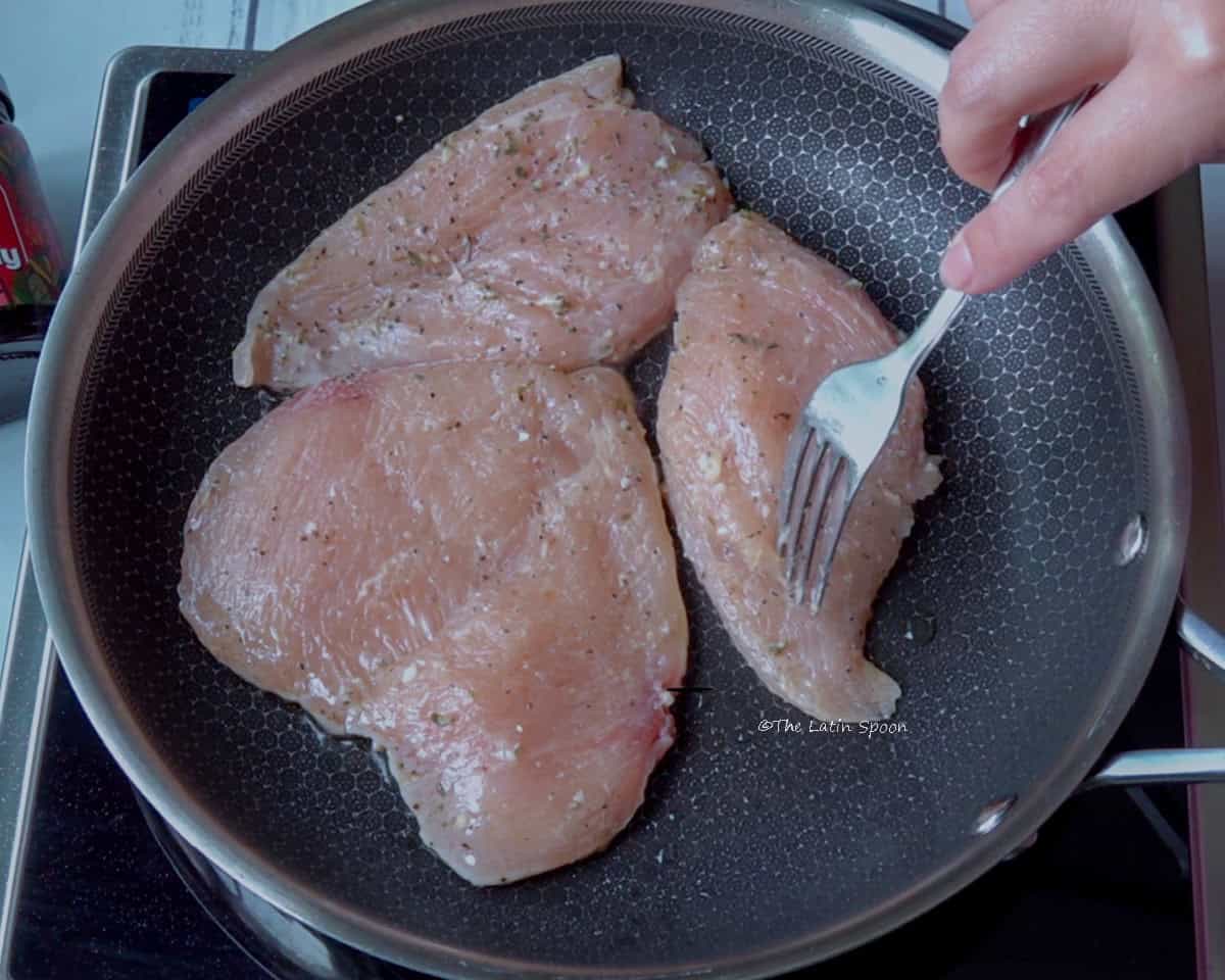 A stainless steel skillet holding 3 pieces of chicken. A right hand is turning the chicken to cook it on the other side.