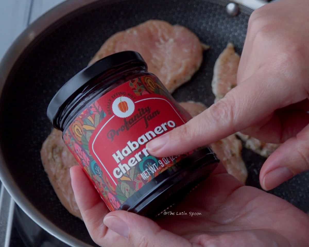 A stainless steel skillet containing 3 pieces of chicken can be seen in the background. Closer to the camera, two hands are holding a jar of jam.