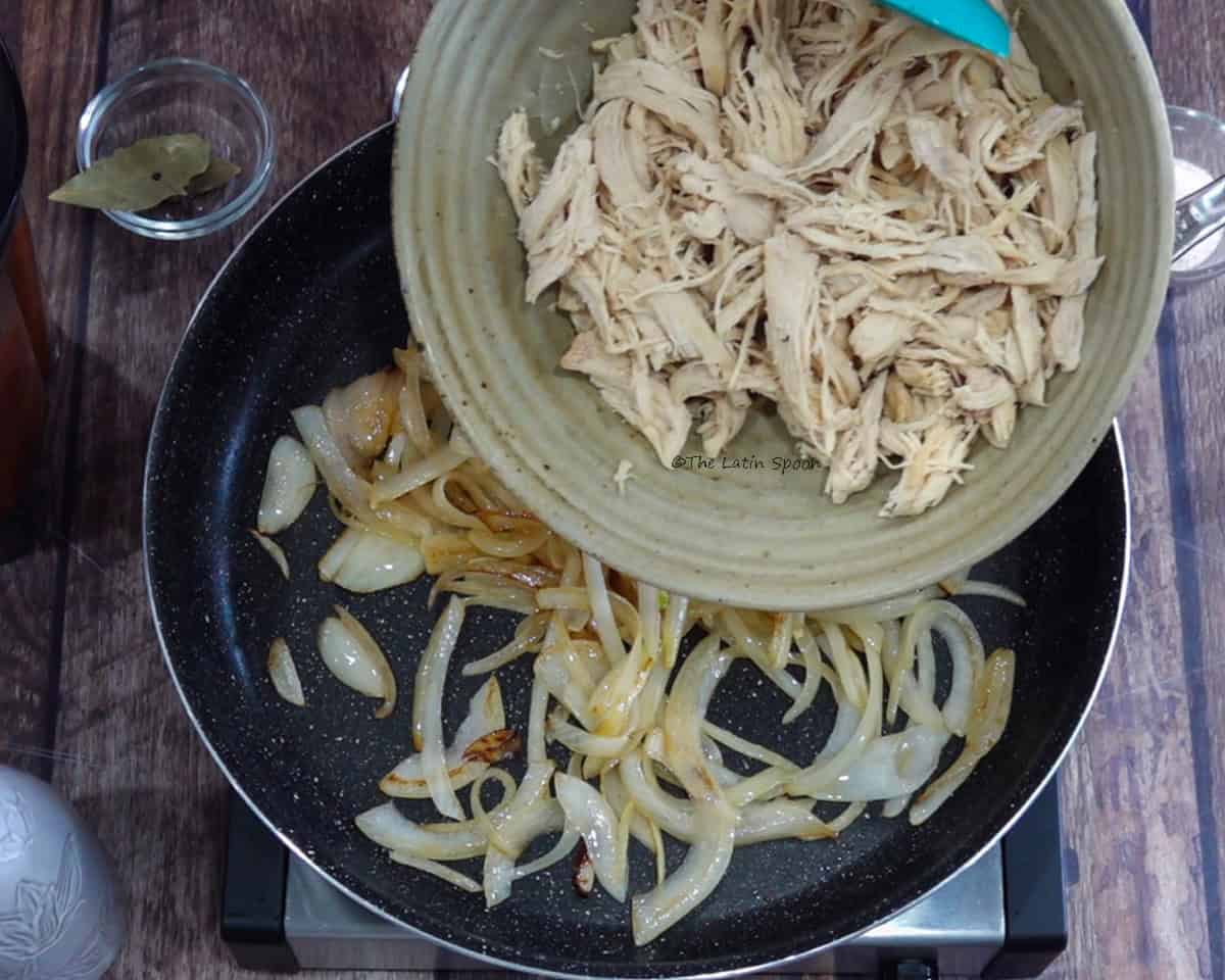 A skillet with caramelized onions while shredded chicken is poured in from a plate above.