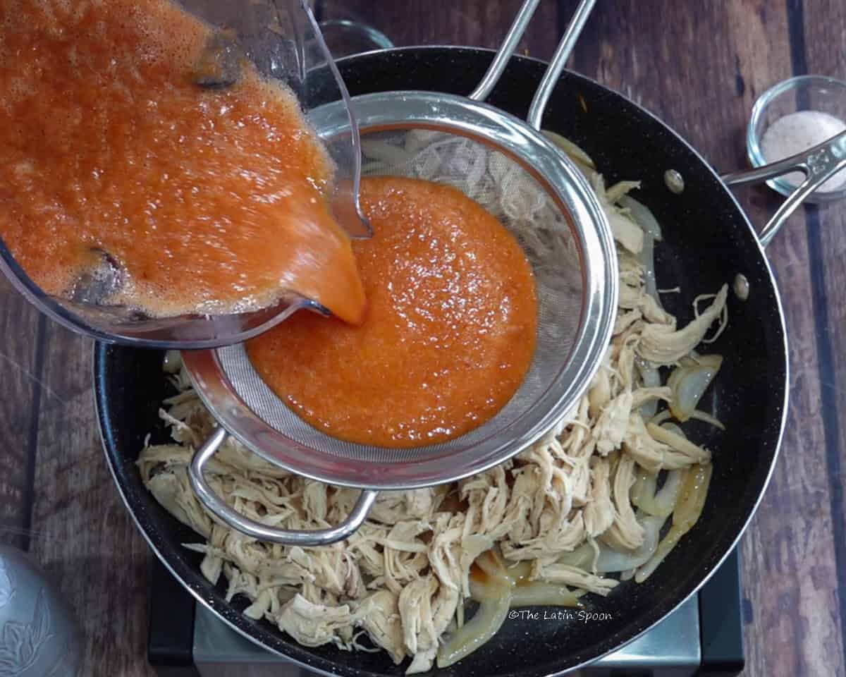 A skillet containing shredded chicken and julienned onions, while tomato sauce is poured from a blender pitcher through a strainer above it.