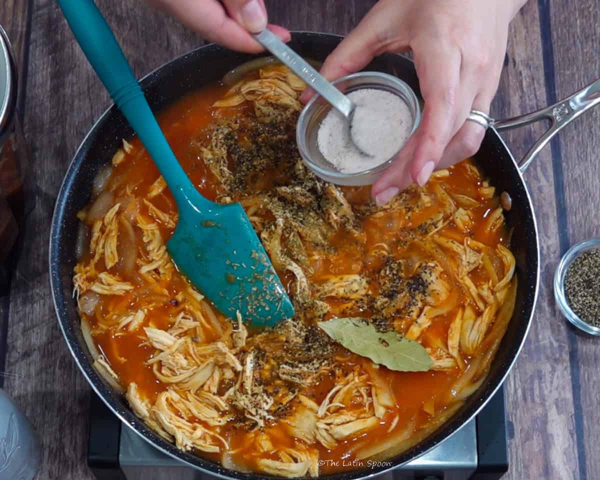 A skillet with shredded chicken in tomato sauce and a spatula inside, with black pepper on top, while one hand holds a small bowl of salt and the other holds a measuring spoon filled with salt above the skillet.