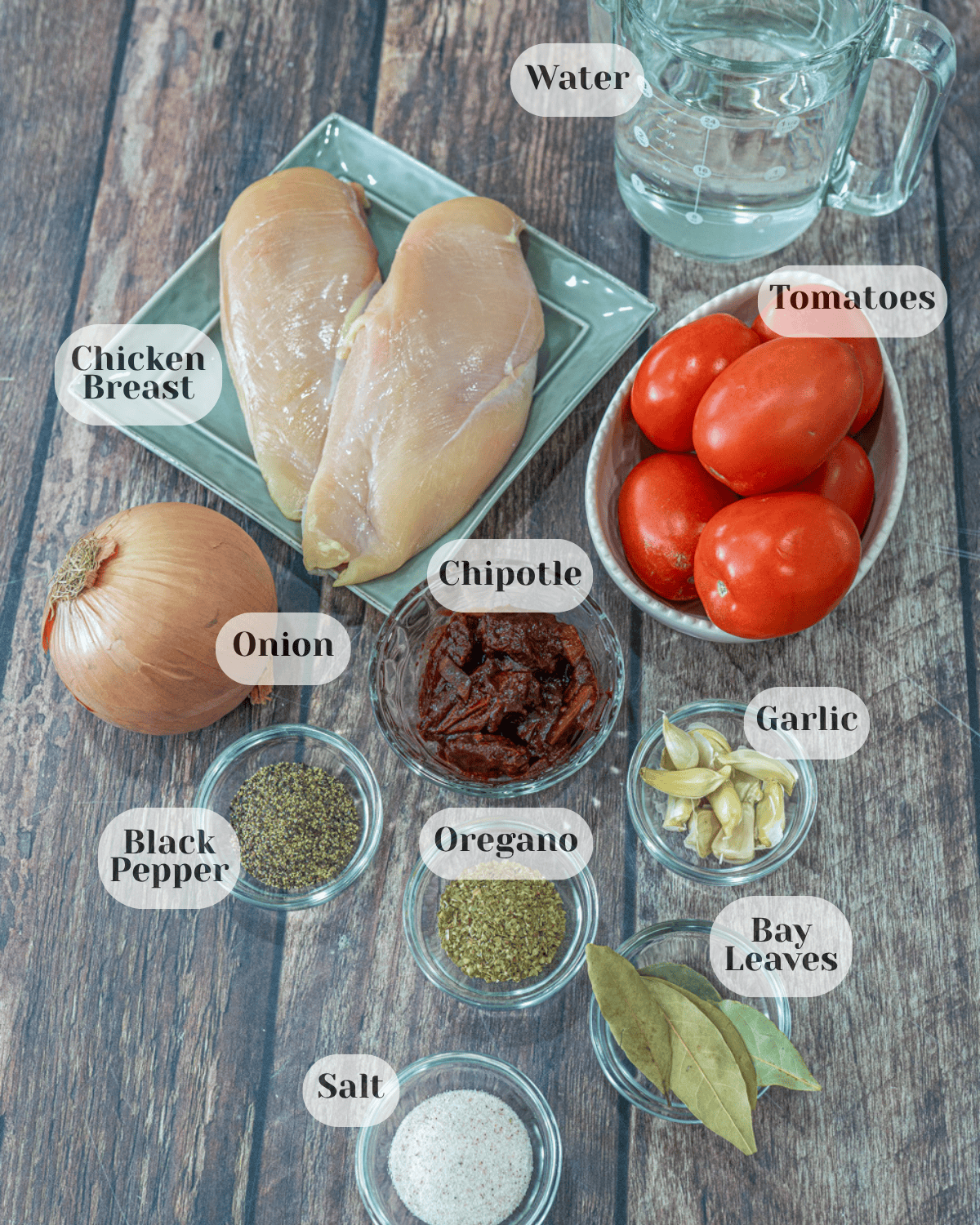 On a countertop, there is a plate with boneless chicken breasts, a pitcher of water, a small bowl of tomatoes, an onion, a small bowl of chipotle peppers in adobo sauce, a small bowl of garlic cloves, black pepper, oregano, bay leaves, and salt.