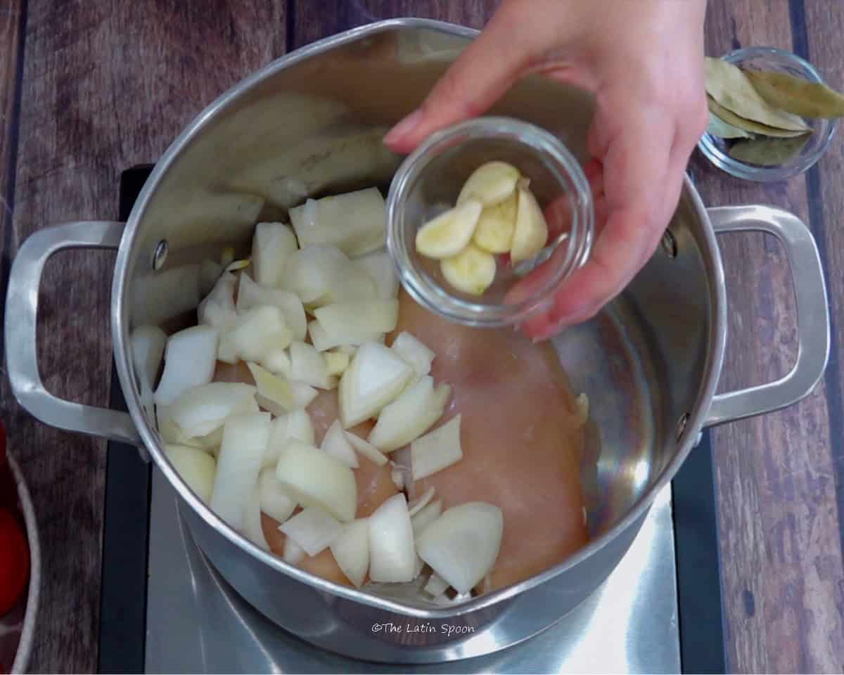 A pot containing chicken breasts topped with pieces of onion, while a hand holds a small bowl of garlic cloves above it and a small bowl of bay leaves sits beside the pot.