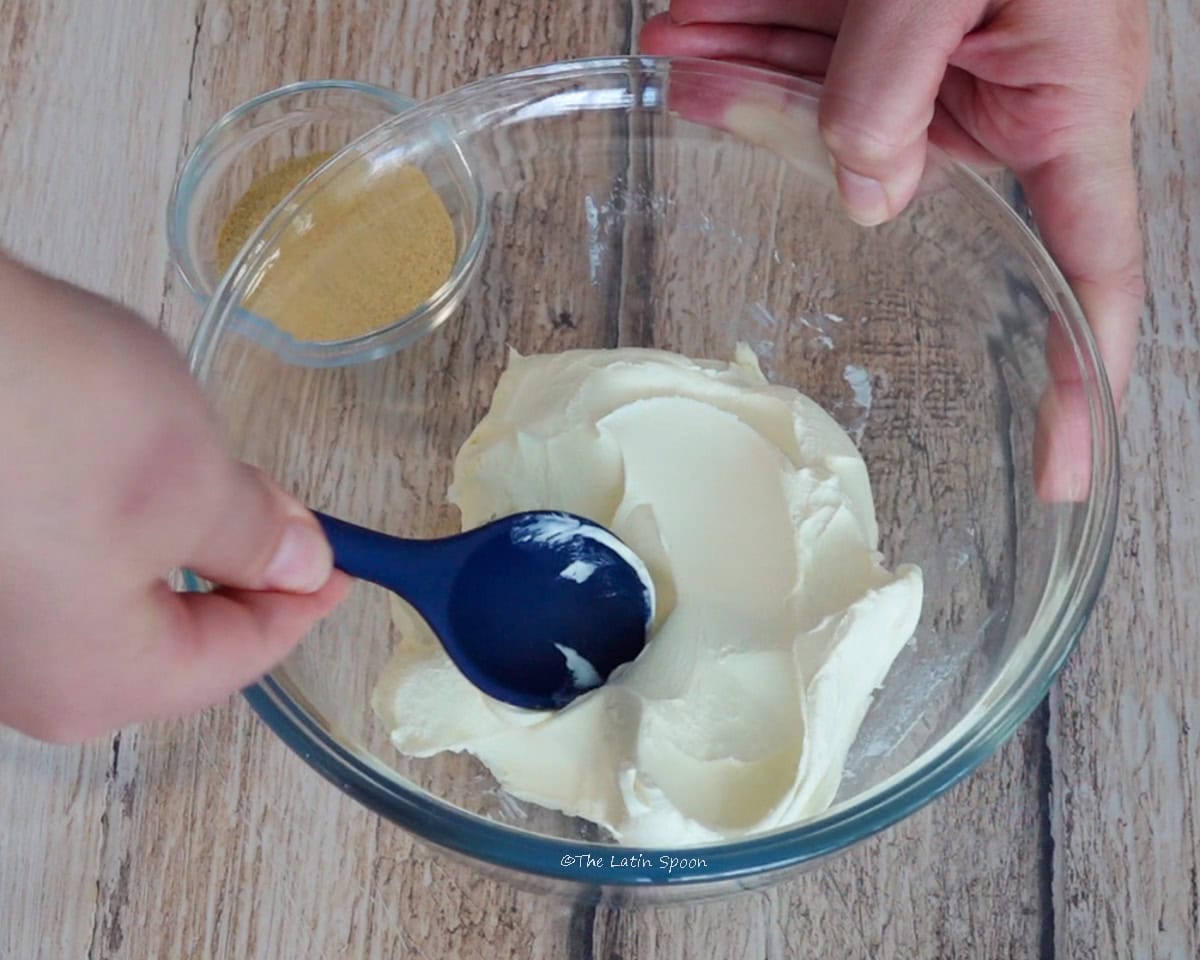 A glass bowl containing cream cheese is held with one hand while the other stirs it with a spoon. A small bowl of garlic powder sits beside it.