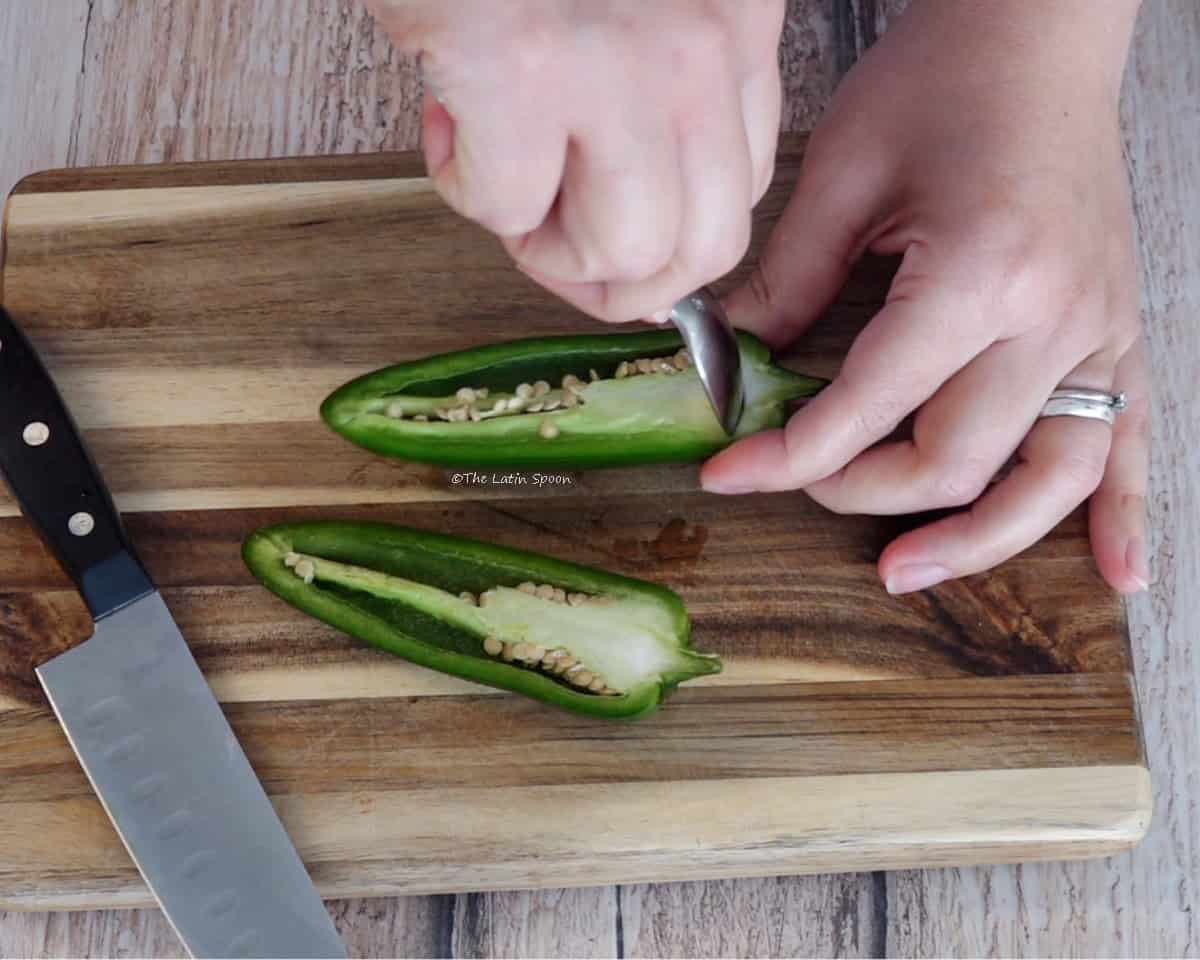On a cutting board, two hands remove the membranes from a jalapeño cut in half. A knife is beside it.