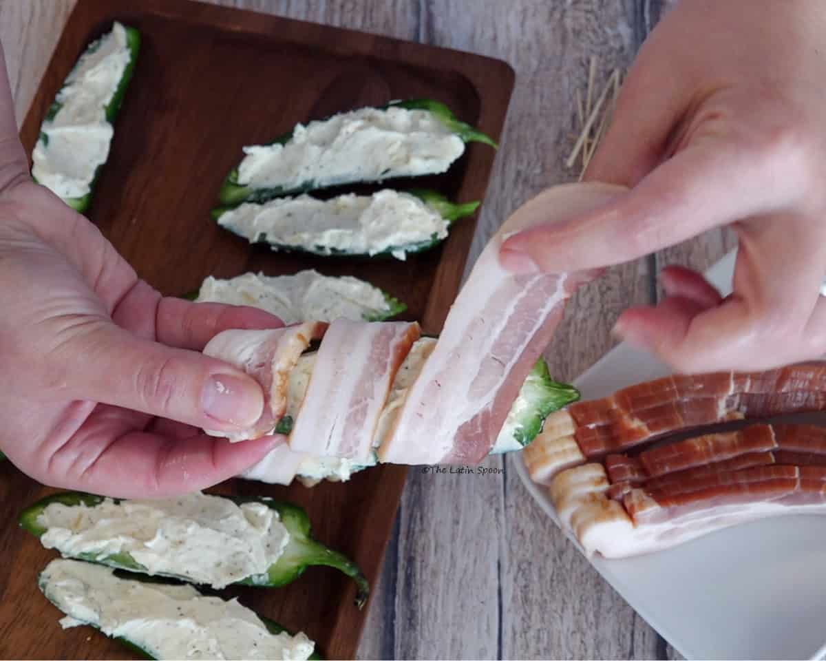 One hand holds a stuffed jalapeño half while the other wraps a strip of bacon around it. In the background, a wooden plate holds the stuffed jalapeño halves, and another plate contains strips of bacon.