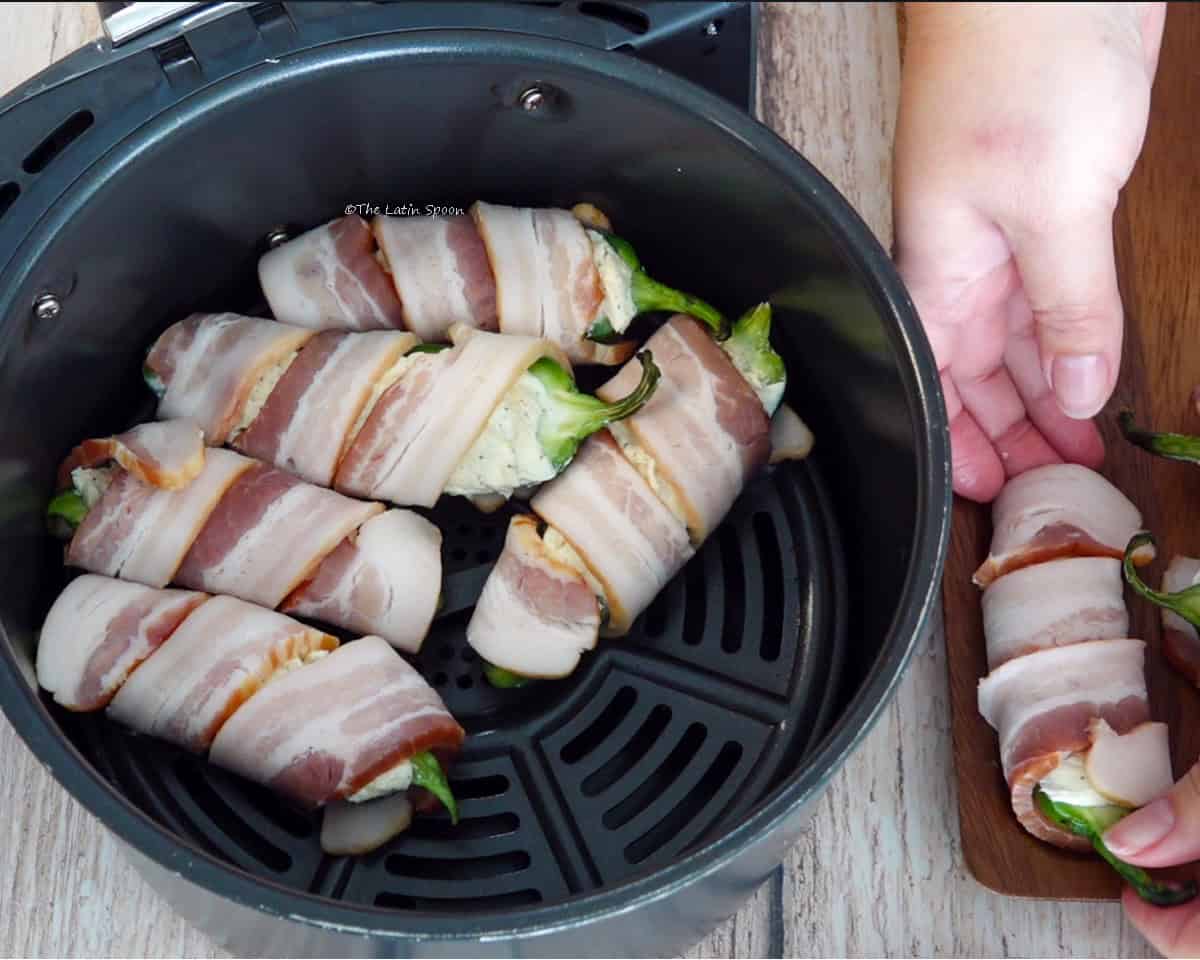 Stuffed jalapeños are inside an air fryer basket while two hands place more jalapeños into the basket.