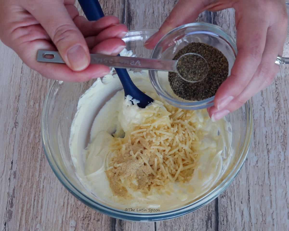 One hand holds a small bowl of black pepper while the other adds it with a measuring spoon to a bowl containing cream cheese and Parmesan cheese. A spatula is inside the bowl.