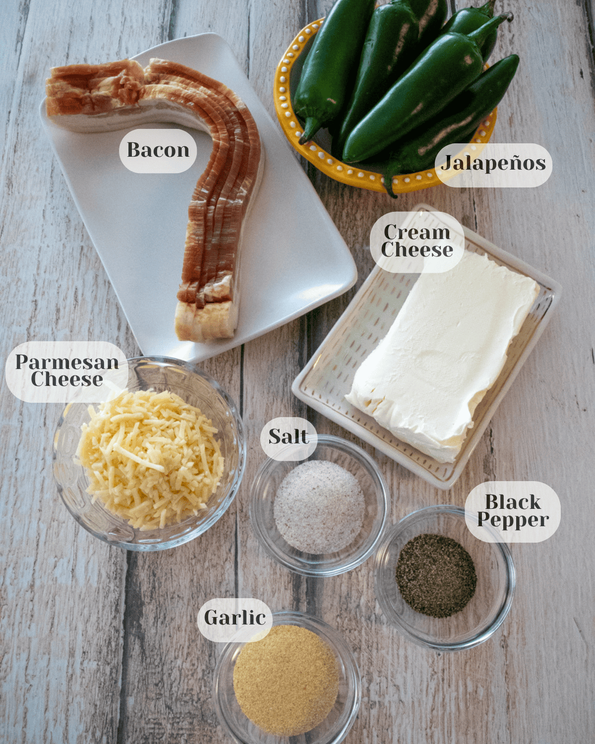 On a countertop, bacon, jalapeños, cream cheese, Parmesan cheese, black pepper, salt, and garlic powder are arranged in separate small bowls.