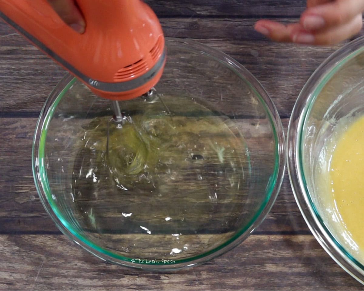 A bowl of egg whites being mixed with a hand mixer, with a bowl of egg mixture beside it.