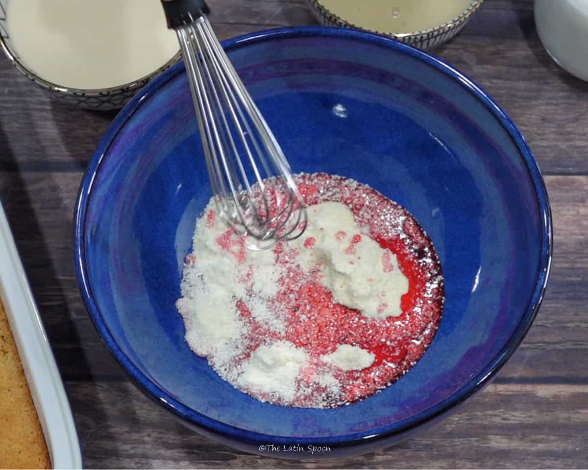 A blue bowl containing a mixture of condensed milk, kola syrup, and powdered milk being mixed with a balloon whisk.