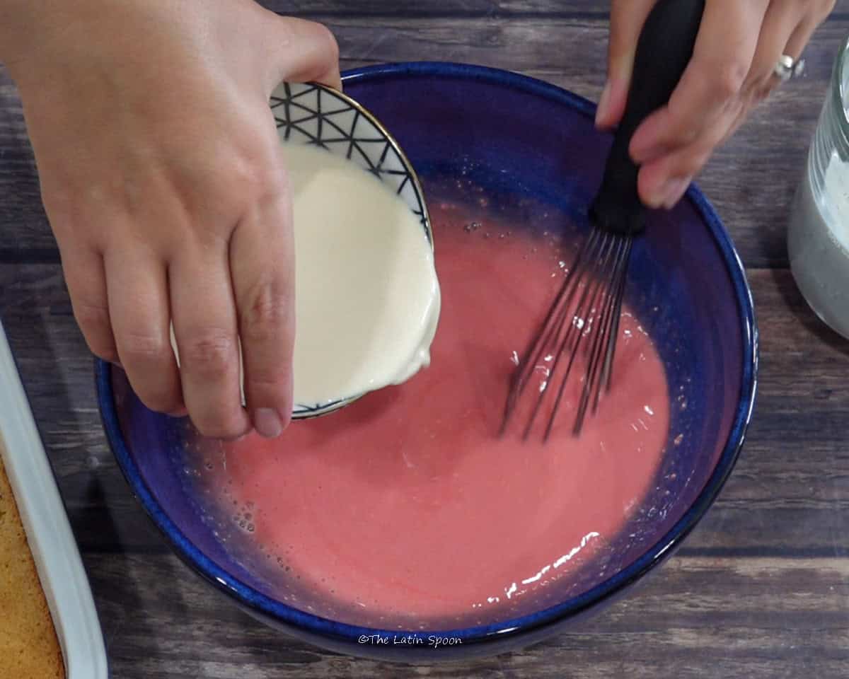 One hand holds a balloon whisk in a bowl containing the milk and syrup mixture, while the other pours milk from a cup into the bowl.