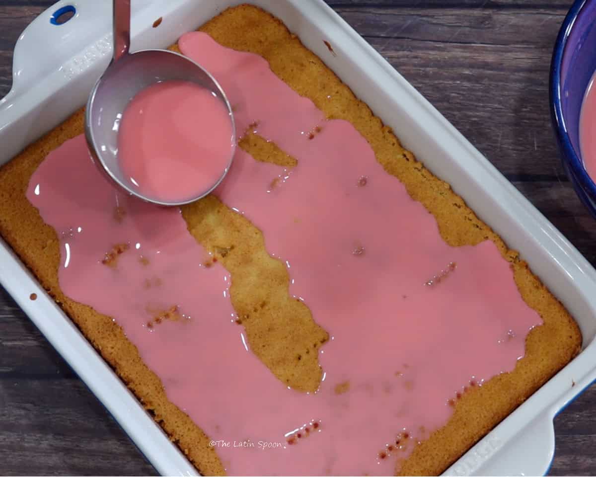 The pierced cake in a glass baking dish being soaked with the milk and syrup mixture using a ladle.