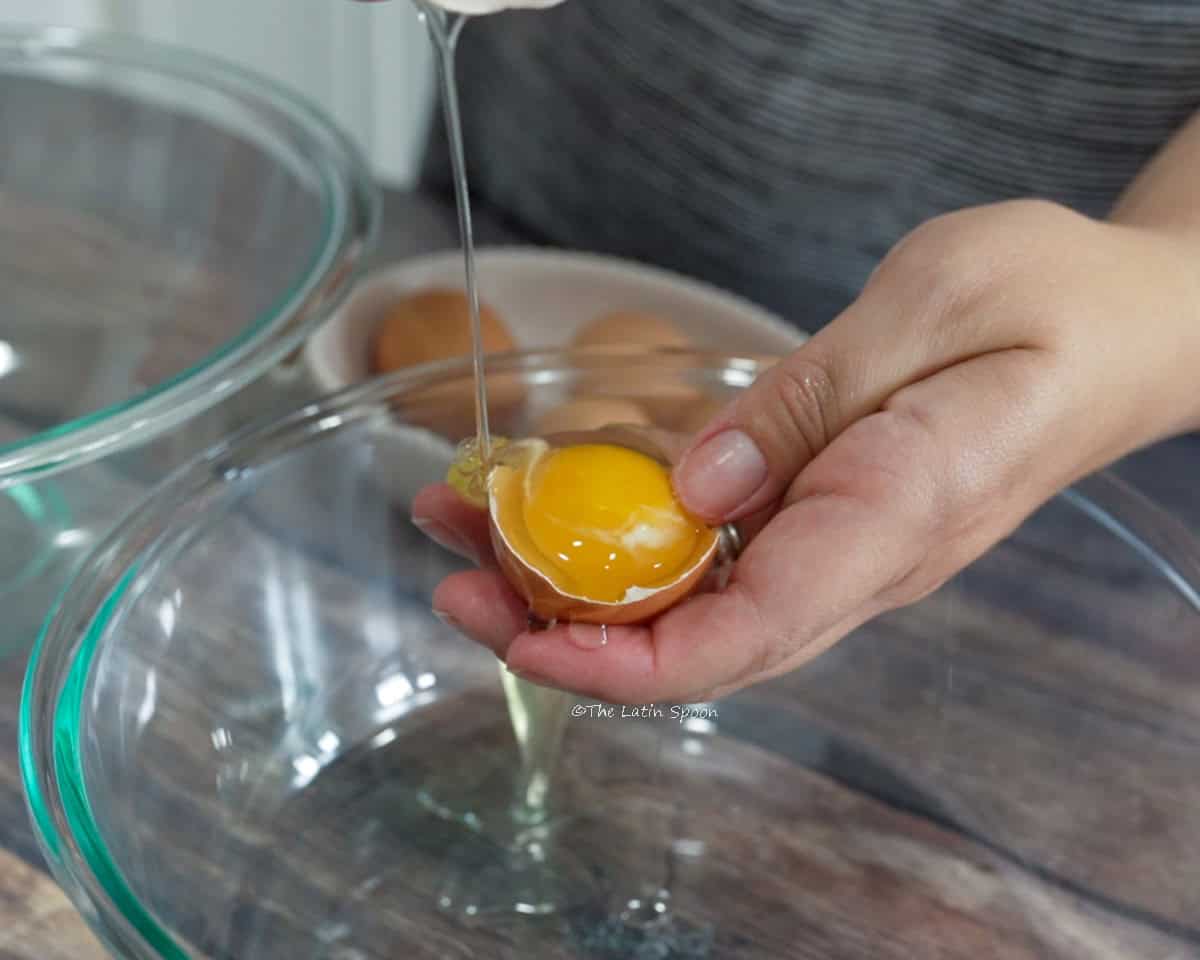 A hand holds an eggshell with the yolk inside while the egg white falls into a bowl.