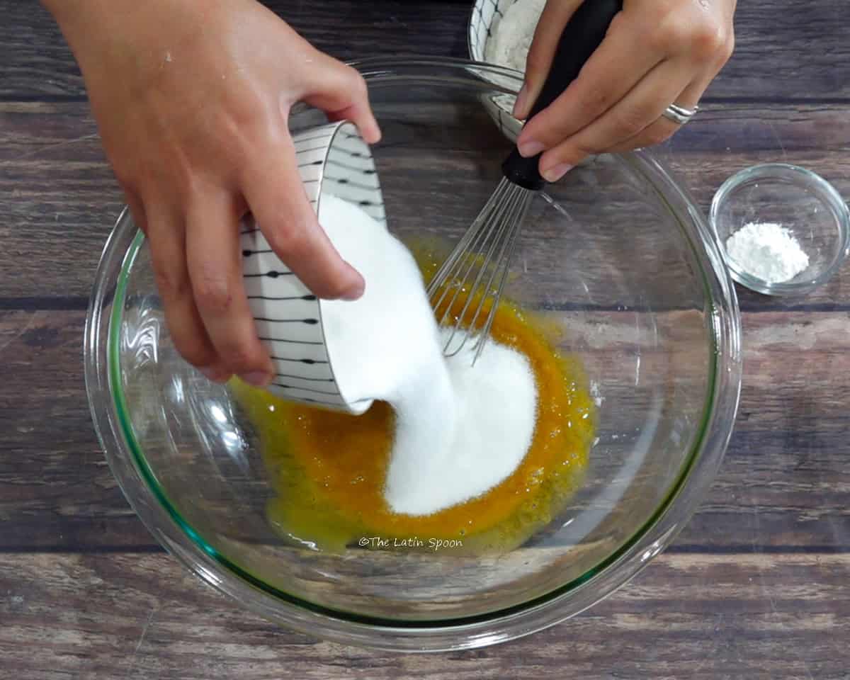One hand pours sugar from a cup into a bowl containing the egg yolk mixture, while the other holds a whisk. A small cup of baking powder is beside the bowl.