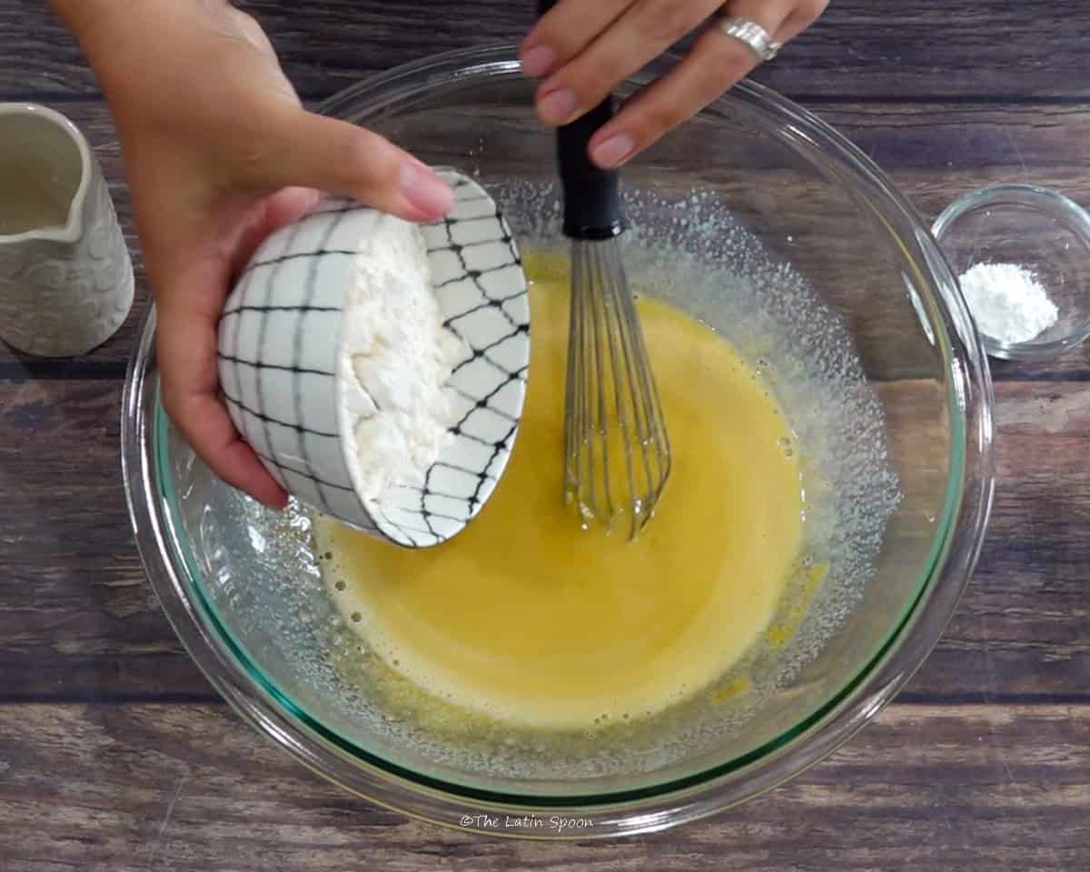 A hand adds flour from a cup to a bowl containing an egg and sugar mixture with a whisk inside.