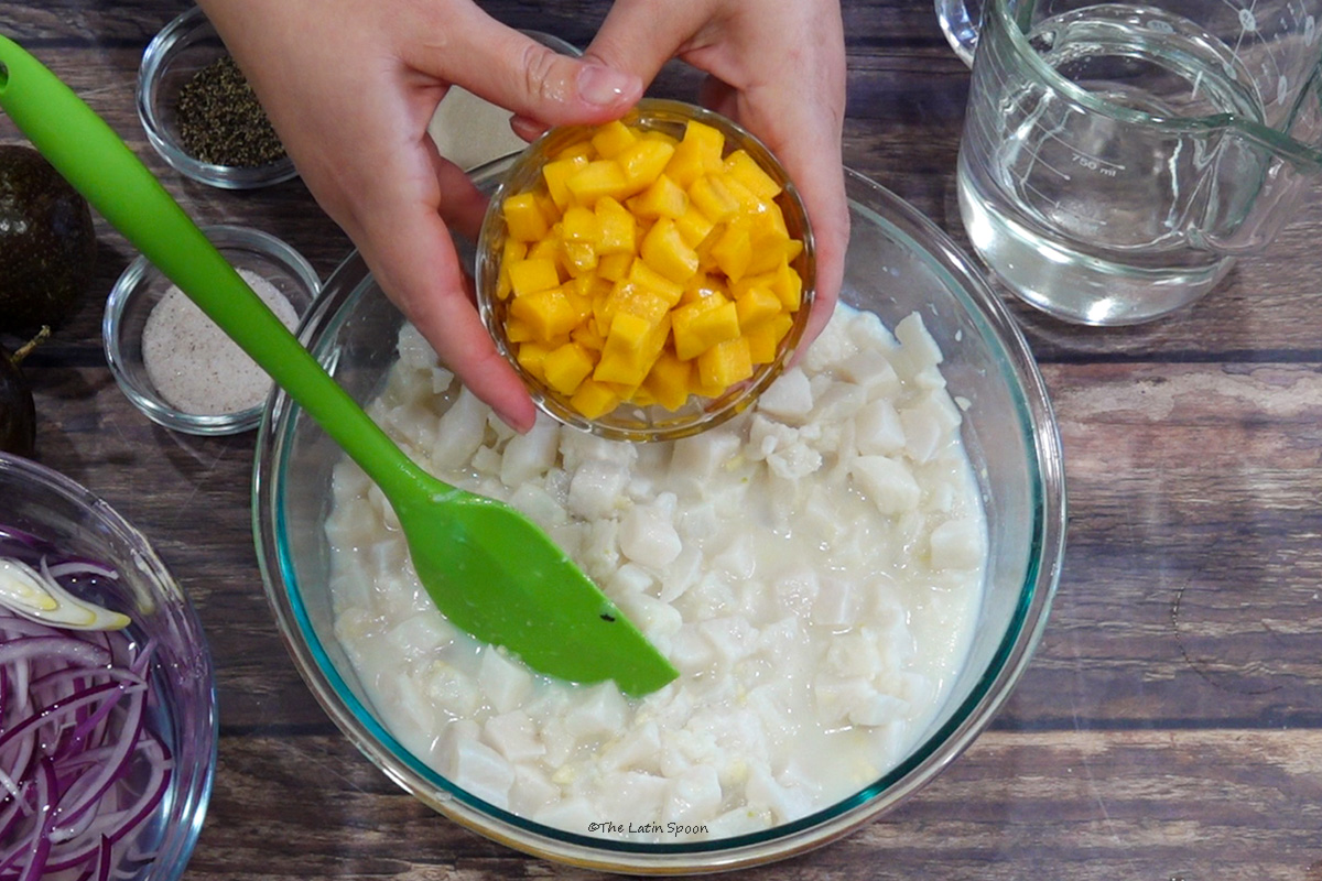 A bowl with a spatula inside. Above it, two hands hold a small bowl of diced mango. Surrounding the bowl are sliced red onion, salt, black pepper, passion fruit, and a measuring cup of Sprite.