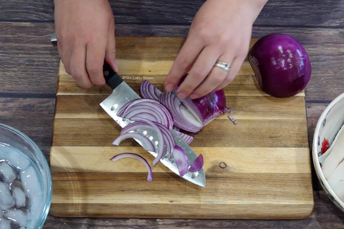 A cutting board with a bowl of ice water beside it. On the board, one hand cuts a red onion with a knife while the other holds the remaining half.