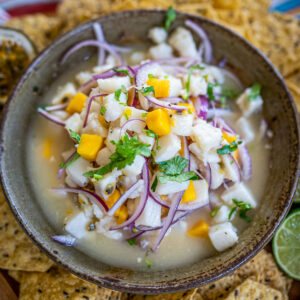 A ceramic bowl filled with tropical ceviche made with mango, red onion, passion fruit, and cilantro. Tortilla chips and lemon wedges are visible in the background.