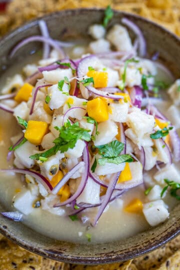 A ceramic bowl filled with tropical ceviche made with mango, red onion, passion fruit, and cilantro. Tortilla chips and lemon wedges are visible in the background.