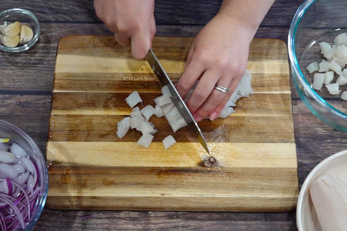 On a cutting board, one hand holds strips of fish while the other cuts across them with a knife to form cubes. Beside the board are a bowl of sliced onions in ice water, several garlic cloves, and a bowl containing cubed fish.