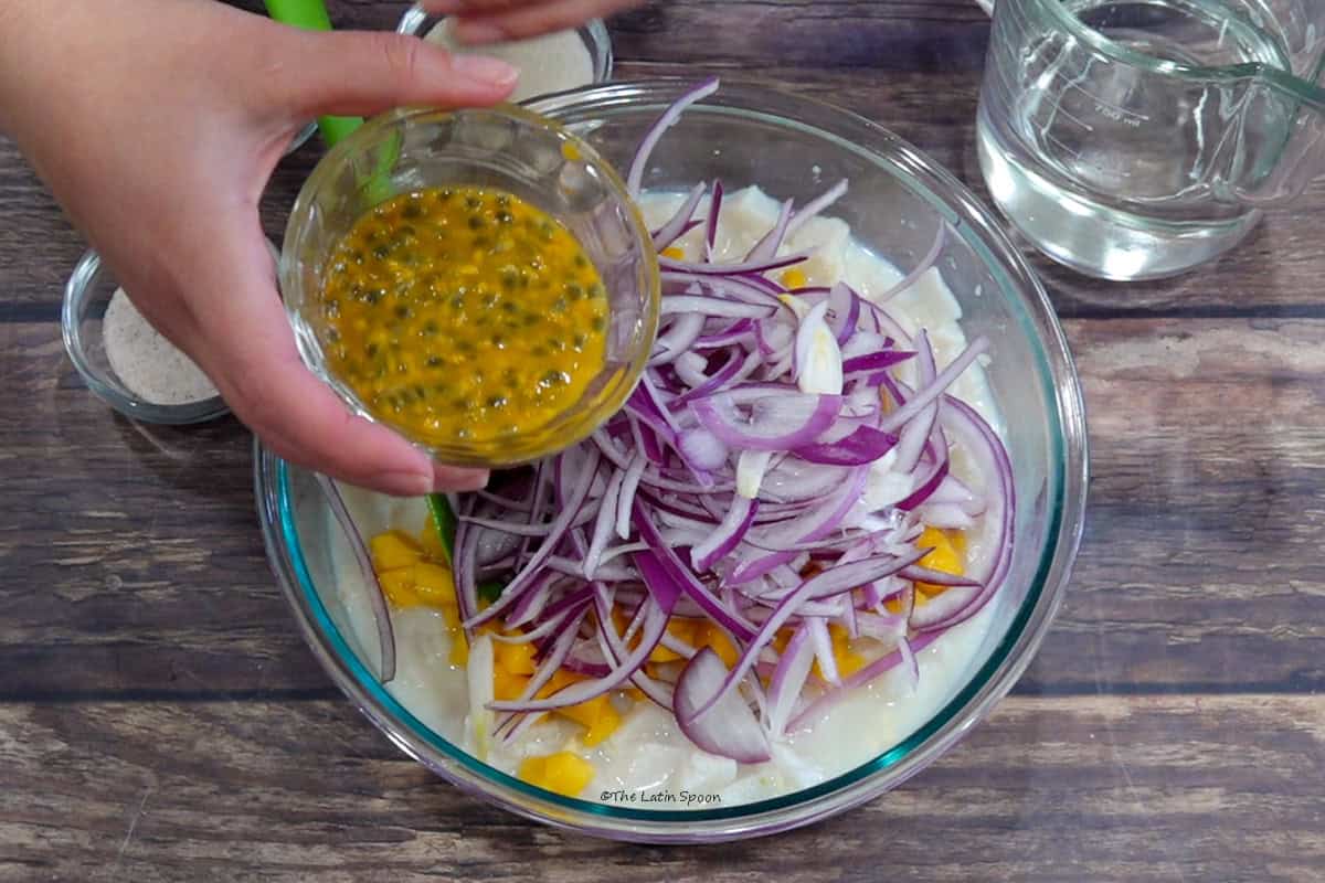 A bowl containing cubed fish, sliced red onion, and diced mango. Above it, one hand holds a small bowl of passion fruit pulp and seeds ready to be added. A measuring cup of Sprite, salt, and sugar are beside the bowl.