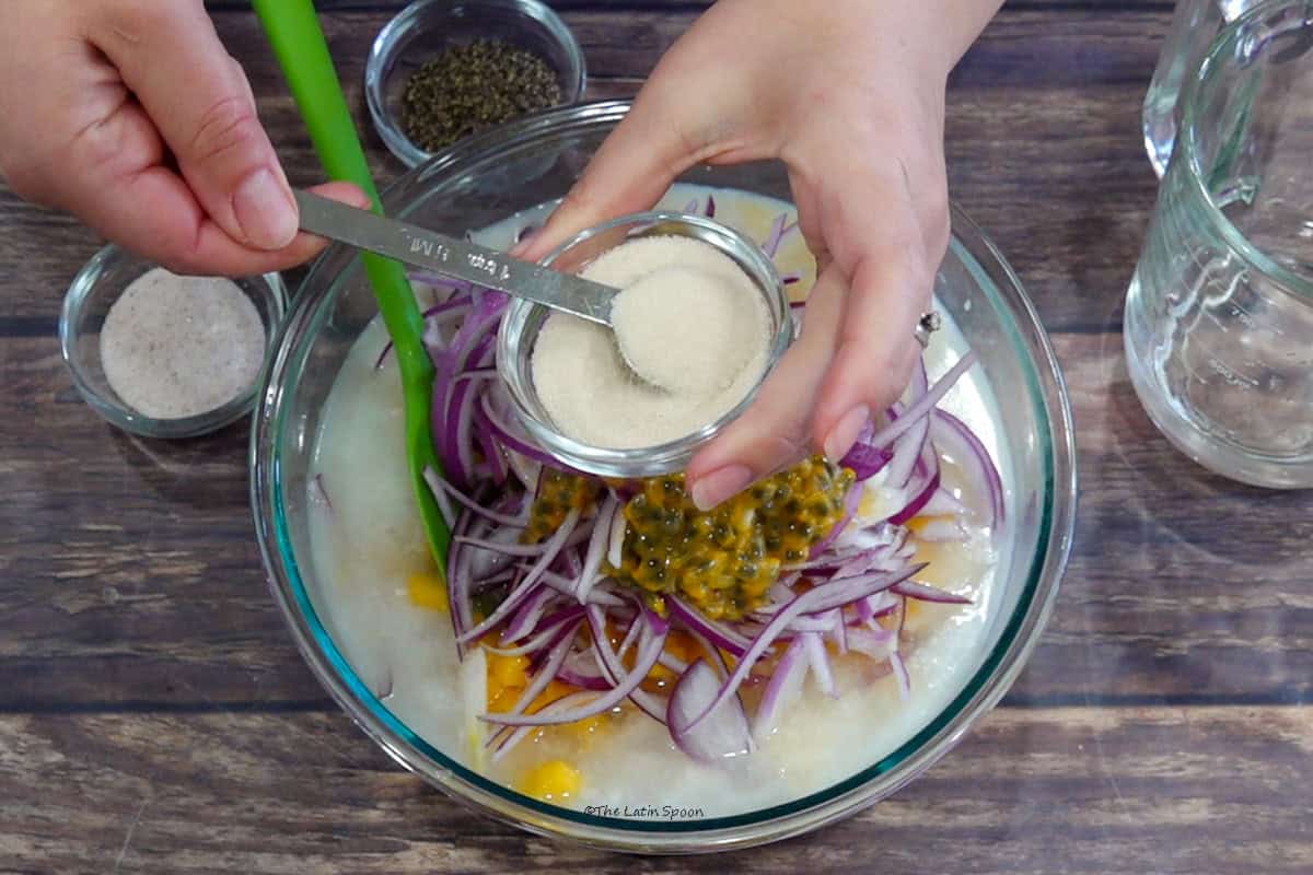 Un tazón con pescado, mango, cebolla y maracuyá; por encima, una tacita con azúcar mientras una cuchara medidora la incorpora. Al lado se ven pimienta, sal y Sprite en una taza de medida.