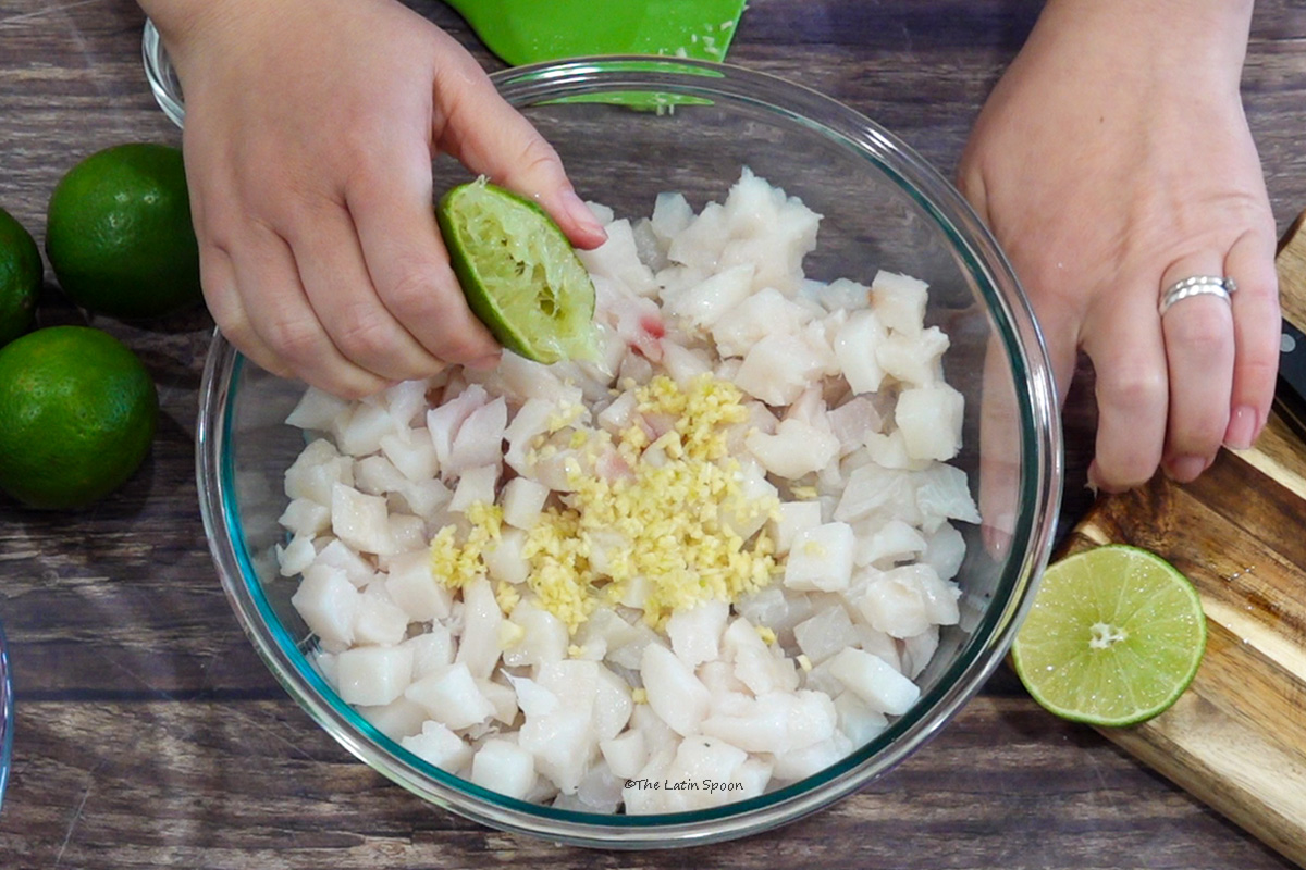One hand squeezes a lime over a bowl containing fish and garlic. Several limes and a spatula are beside the bowl.