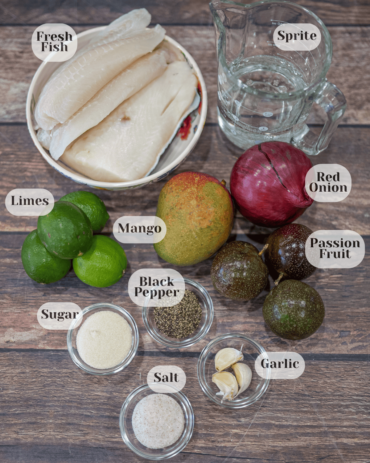 An overhead photo showing the ingredients: fish in a small bowl, Sprite in a measuring cup, one red onion, one mango, three passion fruits, four limes, black pepper in a small bowl, three garlic cloves, sugar, and salt.