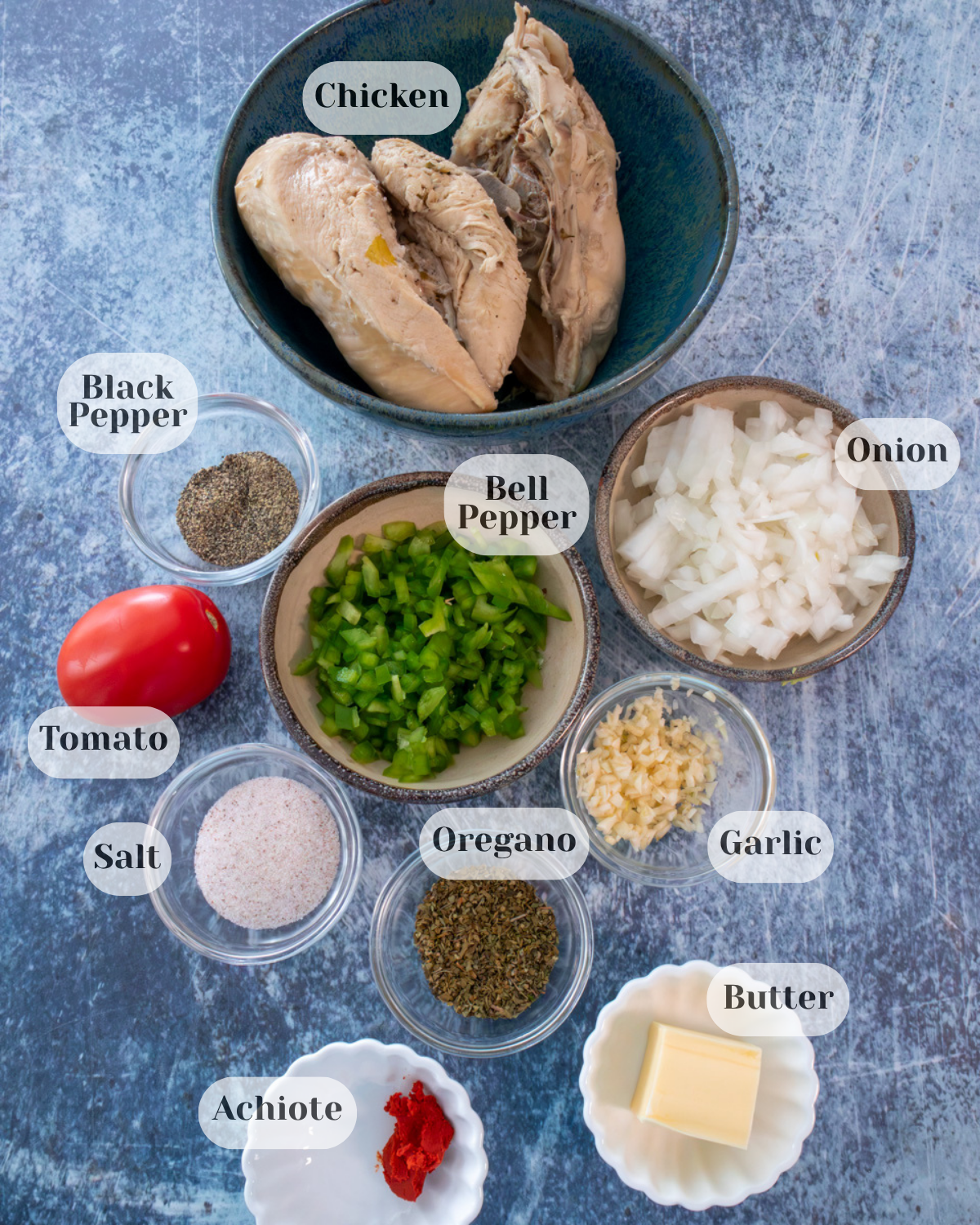 Cooked chicken breasts, black pepper, diced bell pepper, chopped onion, one tomato, minced garlic, salt, oregano, butter, and achiote arranged in small bowls.