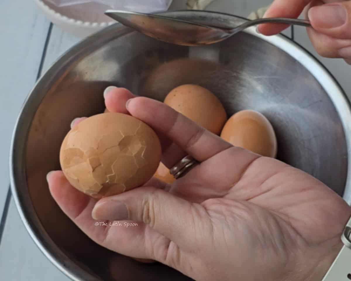 A bowl of cooked eggs while one hand holds an egg above it and taps the shell with a spoon to create a crackled pattern.