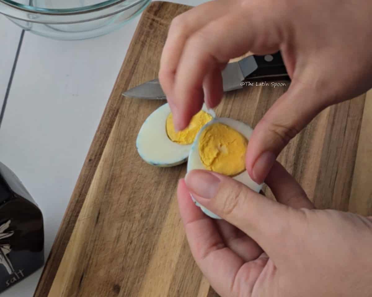 A knife and half of an egg rest on a wooden cutting board while two hands hold the other half and separate the yolk from the egg white.