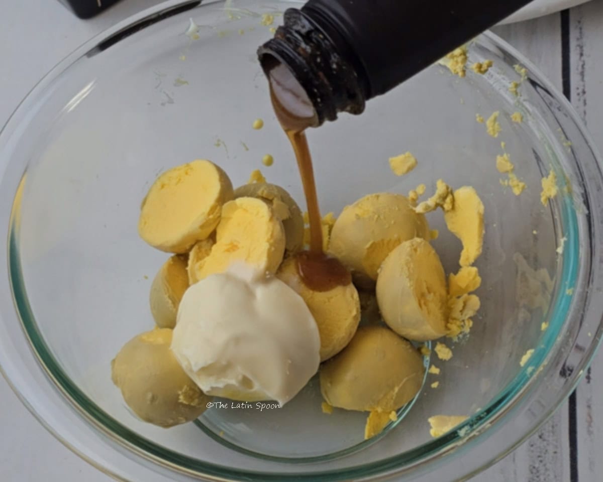 A glass bowl containing the egg yolks and mayonnaise while a small amount of Salsa Lizano is poured into the mixture.
