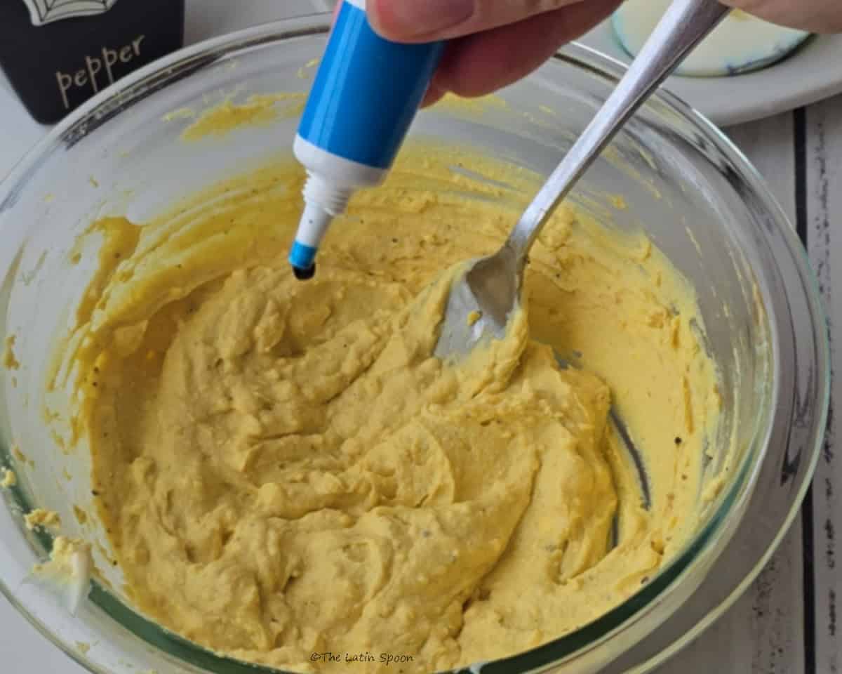 A bowl containing the egg yolk filling with a fork inside while one hand adds a few drops of food coloring from a tube.